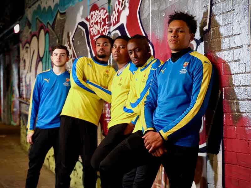 Four people wearing football kit and leaning against a wall covered in graffiti